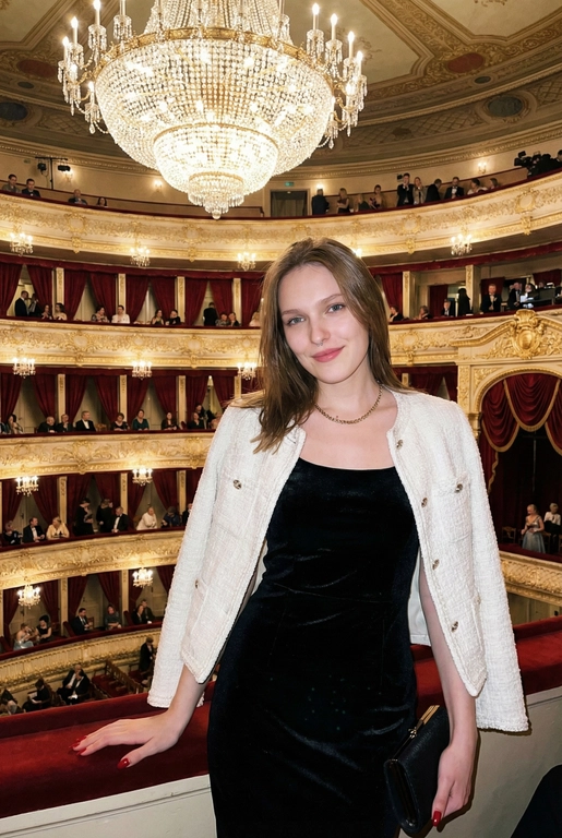 Я стою в ложе Большого театра, на рядов лож и большой хрустальной люстры. В ложах сидят люди, висят красные бархатные занавески. Камера на уровне глаз.  Съёмка выглядит как фото, сделанное на iPhone: естественный свет, лёгкое зерно, минимальная обработка, живой момент без позирования. Взгляд в камеру, легкая улыбка. Голова немного наклонена вбок. Волосы сияют, макияж естественный, губы глянцевые. Одна рука лежит на бортике ложа, во второй - маленькая черная сумочка. Приглушенный свет. Не меняй черты лица девушки с предложенного мной фото. На мне чёрное бархатное вечернее платье без рукавов по фигуре и белый жакет в стиле Channel. Добавь длинные ногти с красным маникюром.