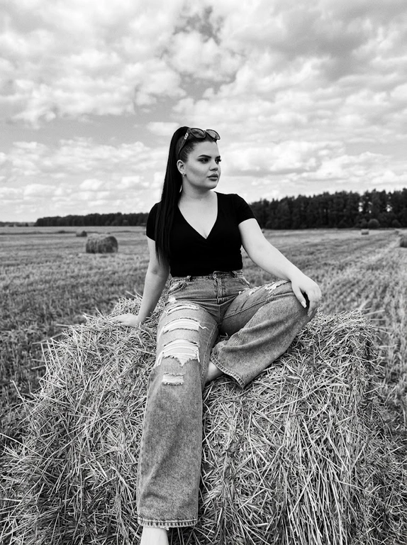 Чёрно-белая high-fashion editorial фотография в сельской местности, женщина с внешностью как на референсе, черты лица не изменять, композиция — более общий план (wide shot), камера на среднем расстоянии, фигура полностью в кадре с окружающим пространством, она сидит на большом тюке сена в поле, корпус слегка развернут, одна нога свободно опущена вниз, вторая согнута, создавая расслабленную, но выразительную позу, руки естественно лежат на колене и на поверхности тюка, взгляд направлен в сторону, профиль, выражение спокойное и уверенное, одежда: чёрный топ с V-образным вырезом и объёмные светлые широкие джинсы с потёртостями и рваными элементами, аксессуары: крупные солнцезащитные очки, минималистичные украшения, фон: открытое поле с несколькими тюками сена на разной глубине, создающими перспективу, линия горизонта ниже центра кадра, чтобы подчеркнуть небо, освещение: жёсткий естественный дневной свет с мягкими облаками, формирует выразительные тени и контраст, стиль: кинематографический, плёночное зерно, высокая детализация, реалистичная текстура кожи и ткани, редакционный fashion стиль, акцент на атмосфере и пространстве кадра


Какая будет прическа?: Гладкие волосы собраны в высокий хвост