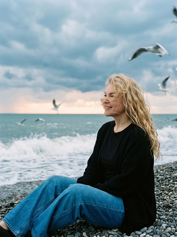 Сохранить внешность 1:1 - лицо, черты, пропорции, возраст, текстуру кожи; фото - единственный эталон. формат 3:4. Атмосферная морская сцена на закате: драматичное облачное небо, холодные голубо-серые оттенки с мягким персиковым у горизонта. Человек сидит боком на гальке у линии прибоя, корпус развернут к морю, взгляд в сторону моря. Ветер растрёпывает волосы, лицо частично перекрыто прядями (волосы развиваются ТОЛЬКО ЕСЛИ они длинные, прическа сохраняется с фото 1:1). в небе летают чайки, часть с лёгким motion blur. Одежда: чёрная базовая футболка, чёрный вязаный кардиган свободной посадки, свободные синие джинсы прямого кроя. Съёмка на винтажную плёночную камеру Kodak, объектив 35mm. Цвет в стиле Kodak Portra 400: матовые тени, мягкий контраст, натуральная цветопередача, лёгкая десатурация, выраженное плёночное зерно. Editorial-композиция, высокий фотореализм, 4K.