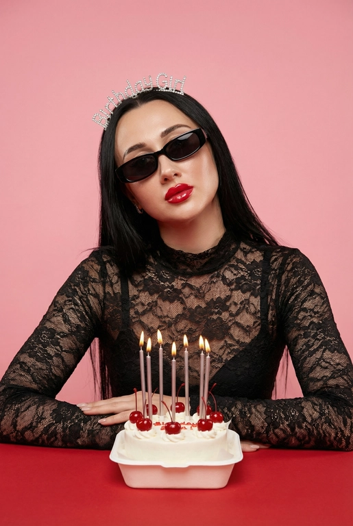 Стильная fashion-фотосессия в студии с розовым фоном. Женщина сидит за красным столом, перед ней белый маленький бенто-торт с вишенками.  На торте тонкие серебристые зажженые свечки. Она в чёрном кружевном топе с длинными рукавами. На голове диадема с надписью “Birthday Girl”, на глазах чёрные узкие солнцезащитные очки. Макияж глянцевый, акцент на губах, чёткий контур, ухоженная кожа.
Женщина слегка наклонила голову в сторону, смотрит в камеру. Настроение кадра — дерзкий день рождения, fashion editorial, ironic glamour. Мягкий студийный свет, ровное освещение без жёстких теней, высокая детализация кожи, чистая композиция. Крупный план / средний план, фокус на лице, без искажений. Профессиональная фотосъёмка, модная журнальная эстетика.