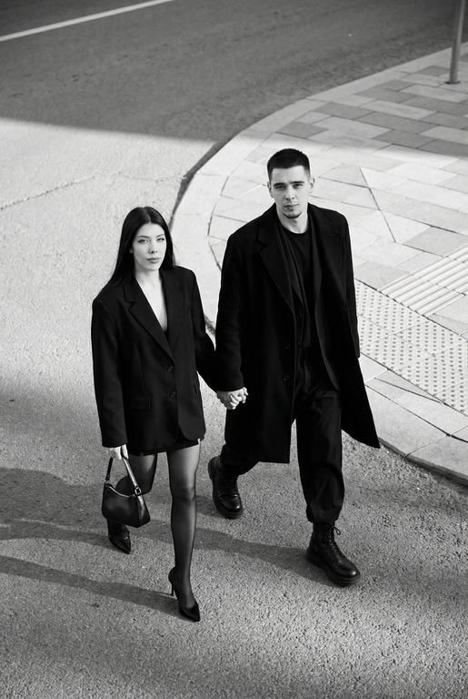 Черно-белая художественная фотография, ультрареализм, 8K, профессиональная съёмка; (оба): в кадре мужчина и женщина; каждый персонаж жёстко привязан к своему загруженному референсу, лица используются напрямую, без генерации, усреднения, подмены или смешения. Вид сверху под углом. Они идут по городской улице, держась за руки; шаг в движении, эффект «пойманного кадра». Смотрим вверх в камеру, выражение спокойное, уверенное, отстранённое.
Полностью чёрные образы modern classic: женщина — оверсайз-пиджак, короткое платье/юбка, плотные колготки, остроносые каблуки, маленькая сумка; мужчина — длинное пальто, свободный костюм, массивные ботинки.
Фон: асфальт, плитка, тактильная дорожка, геометрия.
Контрастный свет, мягкие тени, высокая детализация тканей.
Стиль: black & white, cinematic, high fashion, editorial, minimalism, street photography.
Качество: ultra high resolution, sharp focus, film grain, без ретуши.

