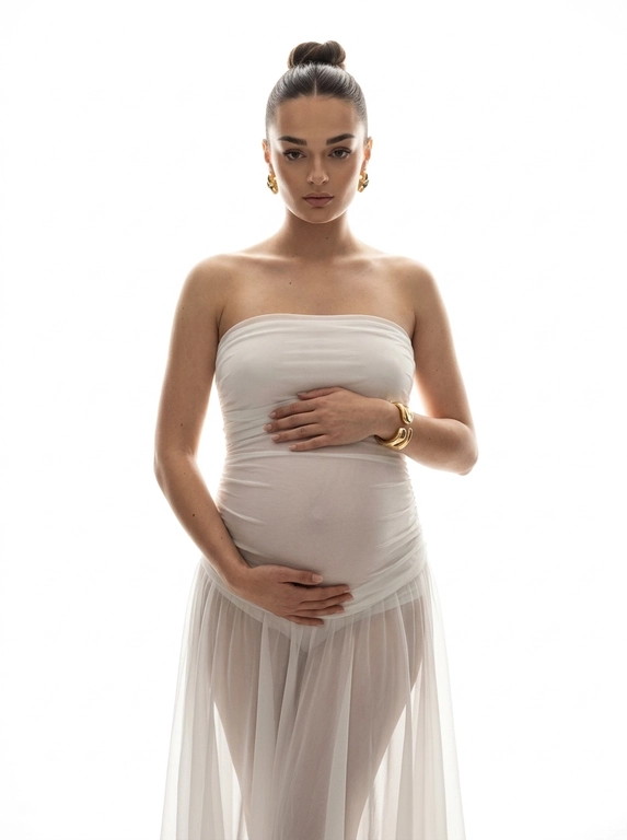 Ultra-realistic high-fashion maternity portrait, framed from knees up. Woman stands centered against a pure white seamless studio background. She wears a strapless sculptural white gown made of sheer layered chiffon and silk, tightly fitted over the pregnant belly and hips, flowing softly downward with translucent folds. The dress emphasizes the round belly clearly and elegantly.

Pose: body slightly angled, both hands gently resting on the belly, one above and one below. Gaze directed straight into the camera, calm, powerful, intimate. Posture upright and confident.

Jewelry minimal: bold gold earrings and a sculptural gold bracelet.

Lighting: high-key studio lighting from behind and slightly from the sides, creating a glowing halo effect around the silhouette, soft rim light outlining the body, gentle frontal fill to keep facial features clear. No harsh shadows, smooth tonal transitions, depth and volume preserved.

Skin looks real and natural with visible texture, no smoothing, no CGI. Editorial luxury maternity aesthetic, timeless, emotional, refined. No text, no logos, no watermark.

Причёска: Hair styled in a sleek high bun, clean and polished, with the hair tightly smoothed back and no loose strands.