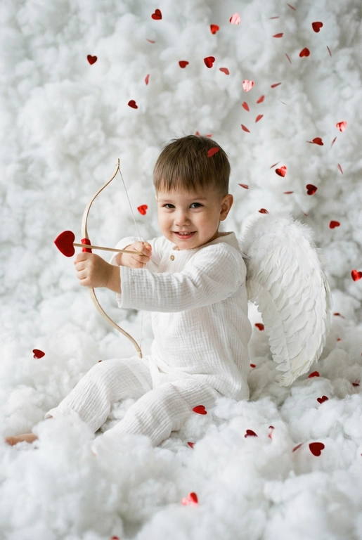 Использовать реальные черты лица человека с референса, естественную текстуру кожи и настоящий цвет и длину волос. Личность должна оставаться неизменной.

Y2K. Сказочная сцена с купидоном, сфотографированная как реальный момент.
Человек сидит среди плотных, пушистых белых облаков, которые полностью заполняют передний, средний и задний план, образуя бесшовную белую среду с многослойной глубиной.

Поза вполоборота: корпус слегка повернут вбок, одно плечо выдвинуто вперед, как будто человек натягивает и отпускает небольшой лук с красной стрелой в форме сердца; руки подняты с легким напряжением, указывающим на движение при выстреле.

Несмотря на поворот тела, голова повернута к объективу, взгляд направлен прямо в камеру. Выражение лица радостное и игривое, с мягкой улыбкой.

Наряд — простая белая пижама: свободная рубашка и брюки из муслина.
На спине — крылья из белых перьев, настоящие и фактурные, с четкими бликами по краям каждого пера.

Вокруг объекта в воздухе хаотично летают только конфетти в форме сердечек — маленькие красные сердечки из глянцевой фольги, расположенные на разном расстоянии: часть в переднем плане, часть растворяется среди облаков.

Без рассеянного света, без мягкого свечения, без студийного глоу.
Текстура кожи реалистичная и детализированная, без сглаживания и без фильтров.
Тонкая зернистость, волшебное, но искреннее ощущение Y2K-редакции — выглядит как фотография, а не визуализация.