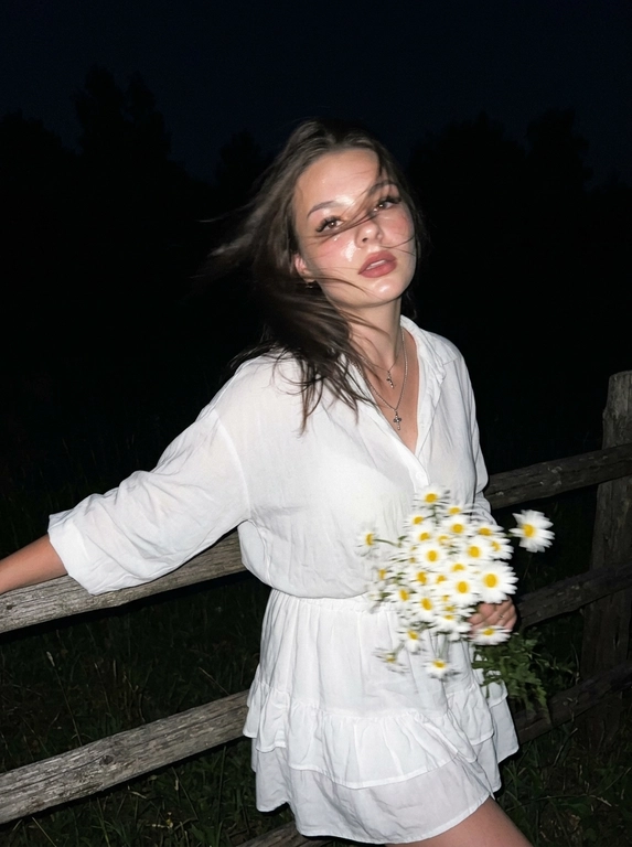 Cinematic candid night shot. iPhone RAW, direct flash, film grain aesthetic.

Girl standing leaning her back and elbows on a wooden fence in a field, distant trees behind her. She suddenly turned her head sharply toward the camera — motion blur from the sharp head turn: hair blur sweeping across the frame, slight blur on face edges, face still recognizable, expression caught mid-movement, alive and unexpected.

hair blown by wind — blur from the sharp turn creates dynamic hair trails across the frame, windswept movement intensifies the motion. Hand holding a bouquet of daisies caught mid-motion.

White short oversized summer blouse with tie detail on chest and ruffles. Matching short skirt with ruffles, same fabric. Beige short suede cowboy boots, white socks slightly visible.

Flawless makeup, dusty rose lips, bronzed tanned skin. Direct camera flash hits face and upper body — skin glowing and shiny, wet oil-skin effect, highlights on cheekbones, nose, forehead. Deep shadows behind her, background falls into dark night.

Dark night, no ambient light except flash. Wooden fence and distant tree silhouettes barely visible in the darkness. Flash creates strong contrast — figure brightly lit, everything behind fades to black.

Cinematic film grain, high ISO sensor noise, muted dark palette, no beauty filter, no plastic skin, no AI look, no watermark, no text.

--ar 9:16 --raw --style motion 50