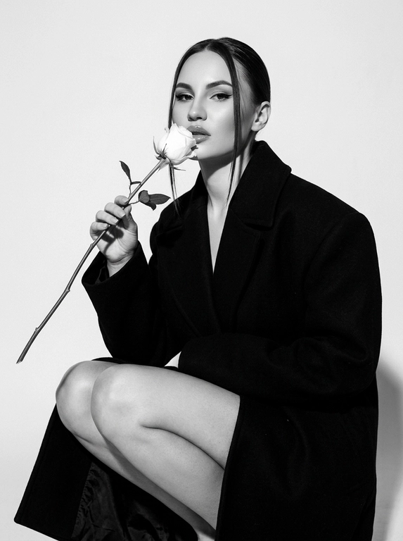 Высоко модная черно-белая редакционная фотография женщины в эффектной позе. Она грациозно присела на корточки, накинув на плечи объемное структурированное черное пальто, оставляя ноги открытыми.
Держит в руке белую розу с длинным стеблем и листьями, поднося к губам бутон. Изящный, минималистичный стиль. Сильное направленное студийное освещение, создающее глубокие тени и высокий контраст. Изысканный макияж с четкой подводкой для глаз и блеском на губах. Чистый фон, эстетика роскошного журнала, стиль Vogue, сверхчеткая, высококонтрастная монохромная фотография. Не изменяйте и не модифицируйте структуру лица, тон кожи или пропорции тела модели.
Только освещение, фон и застывшие текстуры должны быть переосмыслены в контексте задания. Сверхчеткое изображение, высокий динамический диапазон, студийная четкость, реалистичная текстура кожи, естественные поры, едва заметная зернистость пленки, редакционная цветокоррекция, съемка для печати.

Какая будет прическа?: Волосы гладко собранные назад волосы, две прямые пряди спереди выпущены.