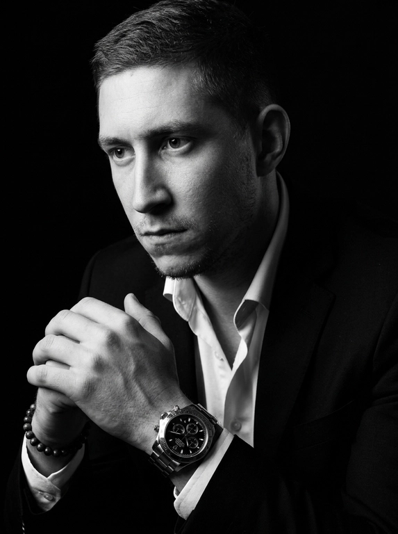 A hyper-realistic 8K cinematic portrait of a uploaded person,
captured in a dramatic Chiaroscuro style. The subject is seated at a
three-quarter angle, leaning slightly forward with a relaxed yet
commanding posture. His face is turned away from the lens, with
one side of his face clearly illuminated while the other is
completely lost in deep, velvety black shadows. His expression is
contemplative, not looking at the camera. The pose is precise: his
hands are clasped together near his chest; one hand is held
straight with fingers slightly tilted, while the other hand is
positioned so only the fingers are visible, resting over the first. He
wears a luxury black chronograph watch with a detailed metal link
bracelet, and a beaded bracelet. His attire is a sharp
black suit jacket over a white dress shirt with the top buttons
open, showing refined fabric textures. The background is a solid,
seamless black. The lighting is sharp and directional, highlighting
the intricate details of his hair,
the moisture in his eyes, and the glint off the watch face.
Cinematic black-and-white color grade with rich contrast and
smooth, natural skin transitions. Shot on 85mm lens, f/1.8, high-
resolution commercial photography