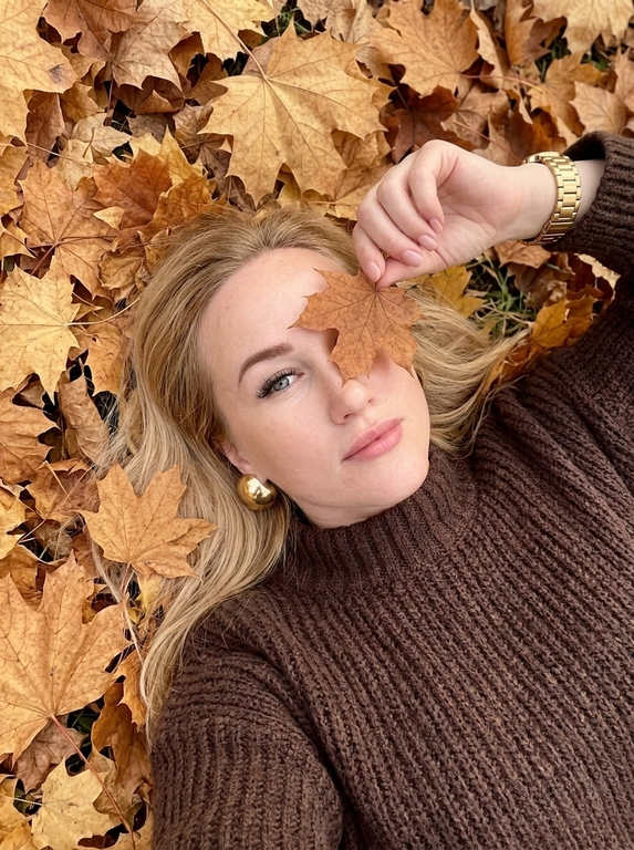 Максимально фотореалистичная фотография, снятая на iPhone. Женщина лежит на спине на плотном ковре из сухих осенних кленовых листьев золотисто-жёлтых, охристых, медных и коричневых оттенков. ракурс: камера находится сверху над женщиной, но не строго вертикально над центром лица — фотограф расположен немного со стороны её корпуса, поэтому кадр идёт сверху вниз под лёгким диагональным углом. Голова находится ближе к верхней части кадра, корпус уходит вниз, лицо и верх тела имеют естественную перспективу. Кадр достаточно близкий — женщина занимает почти всю фотографию, видна примерно от макушки до середины груди, по краям остаётся небольшое количество осенних листьев. Женщина лежит расслабленно, голова немного повернута в сторону, лицо не строго анфас, подбородок слегка развёрнут вбок, при этом глаза смотрят прямо вверх в объектив. Одна рука поднята перед лицом и согнута в локте, женщина тонкими пальцами держит за черенок один сухой коричневый кленовый лист точно напротив одного глаза— лист закрывает один глаз, второй глаз полностью открыт и хорошо виден. На женщине очень объёмный тёмно-коричневый оверсайз свитер из толстой крупной вязки, свободный в плечах и корпусе, с массивным широким высоким горлом и длинными очень широкими рукавами. На поднятой руке поверх рукава свитера находятся одни золотые часы с широким металлическим браслетом, никаких дополнительных часов или браслетов. В ушах крупные эффектные круглые золотые серьги (Не кольца!). Макияж: длинные объёмные пучковые ресницы с 2D-эффектом, кожа с естественной текстурой и видимыми порами, без ретуши. Весь фон состоит только из осенних листьев, без посторонних объектов, слегка пониженная экспозиция, натуральная цветопередача, высокая детализация, фон чёткий без размытия.