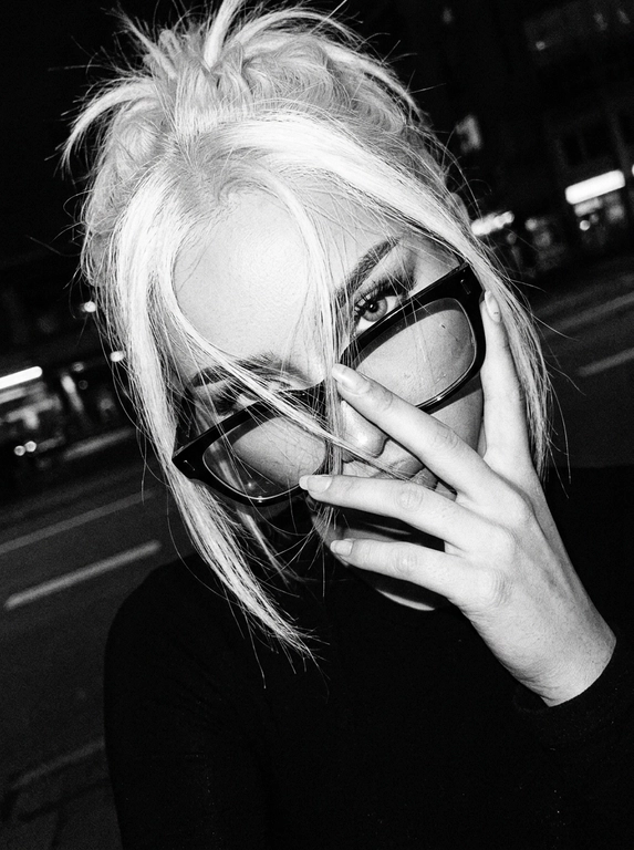 Интригующий, нуарный lifestyle момент; эстетика «messy chic» и ночного города; зернистое черно-белое фото с высоким контрастом, передающее атмосферу скрытности и дерзости.

Крупный план (extreme close-up), голландский угол (наклон камеры); имитация снимка на пленочную камеру 35mm с характерным цифровым шумом и виньеткой.

Модель игриво подносит руку к лицу, частично закрывая его пальцами; кончики пальцев касаются оправы очков или прядей волос.

Динамичный наклон головы в сторону; плечо слегка приподнято, создавая ощущение случайного, небрежного кадра «в моменте».

Загадочный, прямой взгляд в объектив сквозь линзы очков; губы слегка расслаблены; акцент на один глаз, видимый сквозь пряди волос.

Растрепанные волосы; тонкие пряди хаотично спадают на лицо и очки, создавая многослойность и текстуру.

Черный топ; акцент на аксессуары — крупные очки в массивной черной оправе.

Жесткий направленный свет (harsh flash) в темноте, создающий глубокие тени; эффект вспышки «в лоб», который выбеливает кожу и подчеркивает блеск очков и ногтей.

Выраженное пленочное зерно (film grain), легкий расфокус на заднем плане, естественные блики на линзах очков, детальная текстура кожи с сохранением естественных пор под слоем контрастных теней.

Photorealistic cinematic editorial, high-contrast black and white photography, raw aesthetic, 90s grunge vibe.  

Без: ИИ-эффектов, пластиковой кожи, пересветов, HDR, неестественных цветов. Все материалы (волосы, металл, ткань) выглядят максимально достоверно.