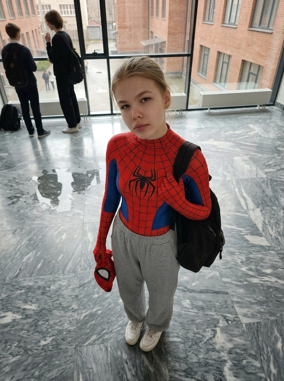 A realistic high-angle full-body photo of a person looking up, at the camera. Keep the same face and facial features from the reference image. Person is wearing a red and blue Spider-Man bodysuit on the upper half, loose grey sweatpants on the lower half, and white sneakers. Person carries a black backpack and holds a red spider-man mask in her left hand. Person is standing on a polished grey marble floor. The lighting is soft and natural, giving a casual cinematic «peter parker after school» vibe.

Доп. характеристики: Ровное лицо，без прыщей