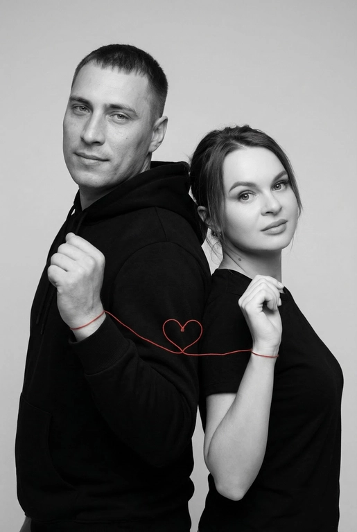 Фотореалистичный чёрно-белый студийный портрет пары, стоящей спина к спине, слегка повернувшись к камере. Оба в минималистичных чёрных образах (худи и футболка), с мягкими уверенными выражениями лица, естественный внешний вид. Тонкая красная нить, связанная вокруг их запястий, соединяет их и образует маленькое сердце между ними. Чистый светло-серый студийный фон, мягкое рассеянное освещение, высокая детализация текстуры кожи, резкий фокус, кинематографичная композиция, вертикальный формат 9:16, минималистичная эстетика, высокое разрешение, профессиональная фотография.