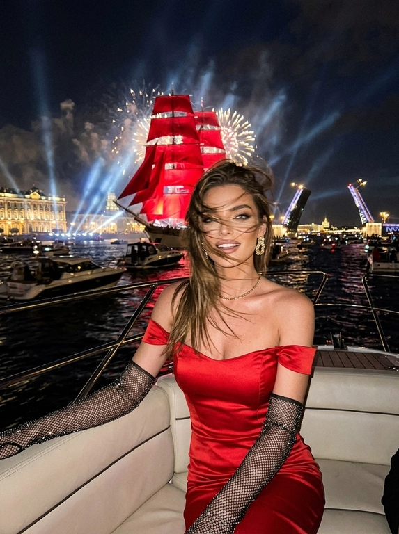 Создай портрет. Ночной экстремально близкий портрет в стиле дорогого fashion-снимка, крупный план, ощущение случайно пойманного момента. Модель, сидящая в энергичной позе на роскошной лодке, плывущей по каналам Санкт-Петербурга во время праздника - "Алые паруса". Волосы развиваются от ветра, слегка на лице. Шикарный вечерний макияж, у нее еле заметная улыбка. 

На заднем плане воды канала, отражающие на горизонте появляются исторические здания, характерные для Санкт-петербурга, рядом с лодкой плывет огромное количество парусных лодок, маленьких яхт, туристических лодок и небольшие лодки вдалеке. Интерьер лодки изысканный. На фоне разводной мост и в центре внимания огромный известный корабль с алыми парусами, паруса подсвечены, с корабля красиво бью прожектора, он в центре внимания, выглядит величественно. Здания подсвечиваются. Все небо залито огромным количеством салютов и фейерверков, а так же светом прожекторов, салюты во весь кадр

Кадр намеренно неидеальный: сильный motion blur, длинная выдержка, высокий ISO, выраженное зерно. Жёсткий контраст, кинематографичная небрежность, ощущение ночи, роскоши и живого, спонтанного кадра. Атмосфера праздника, величественного события. Модель сидит грациозно прямо к кадру, голова повернута в сторону, лицо немного размазано из-за движения, девушка в расфокусе, не смотрит в кадр. Весь фокус на корабле с алыми парусами, они четко подсвечены.

Атласная красная лента "Выпускница 2026"?: Без ленты

Какое будет платье?: Она одета в черное очень приталенное атласное черное платье ниже колена с открытыми плечами, на ней длинные перчатки из сетки со стразами; золотые сверкающие серьги, тонкая золотая цепочка-ожерелье.