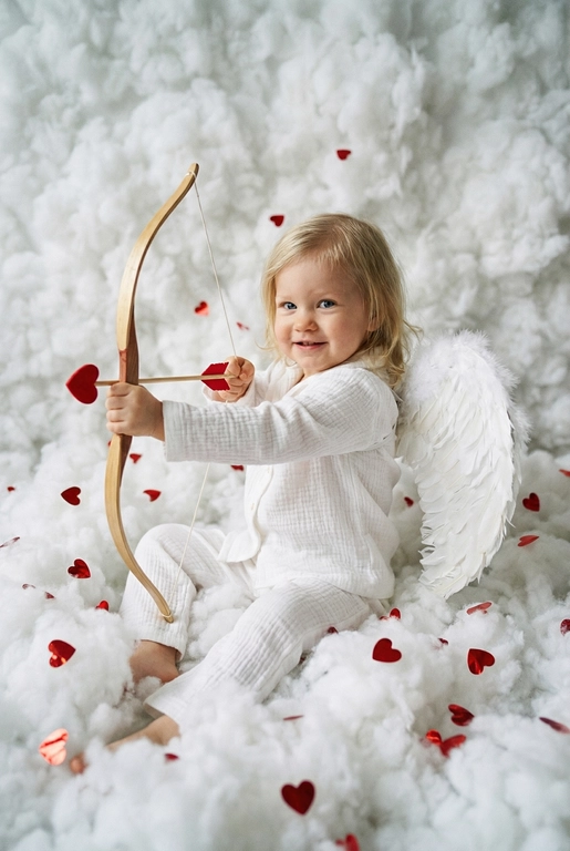 Использовать реальные черты лица человека с референса, естественную текстуру кожи и настоящий цвет и длину волос. Личность должна оставаться неизменной.

Y2K. Сказочная сцена с купидоном, сфотографированная как реальный момент.
Человек сидит среди плотных, пушистых белых облаков, которые полностью заполняют передний, средний и задний план, образуя бесшовную белую среду с многослойной глубиной.

Поза вполоборота: корпус слегка повернут вбок, одно плечо выдвинуто вперед, как будто человек натягивает и отпускает небольшой лук с красной стрелой в форме сердца; руки подняты с легким напряжением, указывающим на движение при выстреле.

Несмотря на поворот тела, голова повернута к объективу, взгляд направлен прямо в камеру. Выражение лица радостное и игривое, с мягкой улыбкой.

Наряд — простая белая пижама: свободная рубашка и брюки из муслина.
На спине — крылья из белых перьев, настоящие и фактурные, с четкими бликами по краям каждого пера.

Вокруг объекта в воздухе хаотично летают только конфетти в форме сердечек — маленькие красные сердечки из глянцевой фольги, расположенные на разном расстоянии: часть в переднем плане, часть растворяется среди облаков.

Без рассеянного света, без мягкого свечения, без студийного глоу.
Текстура кожи реалистичная и детализированная, без сглаживания и без фильтров.
Тонкая зернистость, волшебное, но искреннее ощущение Y2K-редакции — выглядит как фотография, а не визуализация.