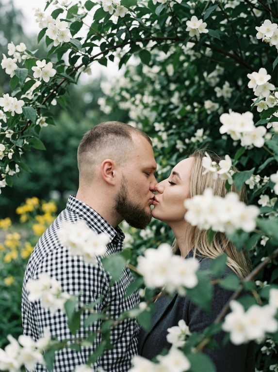 A man and a woman stand face to face, kissing—their lips touching, faces in profile, eyes closed. The shot is close-up, from shoulders to head, angled at eye level, with their faces centered in the frame and surrounded by flowering branches. The foreground and edges of the frame are filled with out-of-focus branches and flowers. Do not alter the faces from the uploaded photos.
The skin appears perfectly smooth and matte.
The man is wearing a cotton shirt with a small gingham check in black and white tones. The woman is wearing a dark gray jacket.
The location is a flowering tree with dense branches: large white flowers with five petals and yellow stamens —presumably jasmine or bird cherry—and dense dark green foliage. The branches frame the figures on all sides, creating a living, arched tunnel. In the left middle ground, yellow flowers are out of focus.
The lighting is natural, overcast, diffused, with no clear direction of the light source—an even, soft light. Shadows are virtually absent. Contrast is low. The overall tone of the frame is muted and cool, with green and white dominant tones. The highlights on the skin are subdued and matte. The film grain is pronounced.
Shot with an 85mm lens, minimal depth of field with sharp focus exclusively on the lips and the area where the faces meet—the foreground with the man's shoulder and the nearby branches are completely out of focus, the background with foliage is blurred into bokeh. Pronounced film grain. A reportage paired shot in the style of a Candid Film portrait, highly detailed and photorealistic.
Soft romantic spring botanical mood,
dominant tones: white blossom, deep green, dark charcoal,
low saturation with cool green emphasis,
flat diffused overcast natural lighting,
minimal shadow definition,
heavy analog film grain throughout,
lifted blacks with a soft hazy quality,
shallow depth of field with strong foreground blur,
Candid Film Spring color grading.