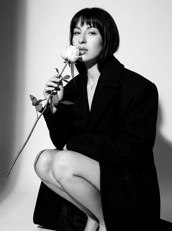 Высоко модная черно-белая редакционная фотография женщины в эффектной позе. Она грациозно присела на корточки, накинув на плечи объемное структурированное черное пальто, оставляя ноги открытыми.
Держит в руке белую розу с длинным стеблем и листьями, поднося к губам бутон. Изящный, минималистичный стиль. Сильное направленное студийное освещение, создающее глубокие тени и высокий контраст. Изысканный макияж с четкой подводкой для глаз и блеском на губах. Чистый фон, эстетика роскошного журнала, стиль Vogue, сверхчеткая, высококонтрастная монохромная фотография. Не изменяйте и не модифицируйте структуру лица, тон кожи или пропорции тела модели.
Только освещение, фон и застывшие текстуры должны быть переосмыслены в контексте задания. Сверхчеткое изображение, высокий динамический диапазон, студийная четкость, реалистичная текстура кожи, естественные поры, едва заметная зернистость пленки, редакционная цветокоррекция, съемка для печати.

Какая будет прическа?: Не изменять мою прическу.