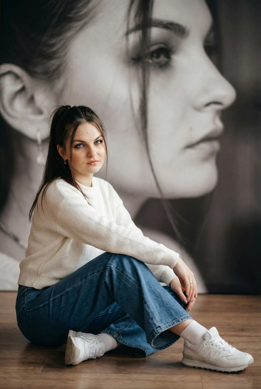   Максимально сохранить внешность, причёску и длину, как на фото.
Женщина должна сидеть на полу в изящной позе, в современном белом джемпере , в широких джинсах и белых носках и белых  кроссовках New Balance.

Её волосы как на фото
Выражение лица, как на фото, она смотрит на камеру .

Фон — тот же её профиль, в мягком крупном чёрно-белом художественном стиле, крупный план сбоку.

Композиция должна быть художественной, мягкой, реалистичной.
Создай изображение, которое выглядит как настоящая фотография.
