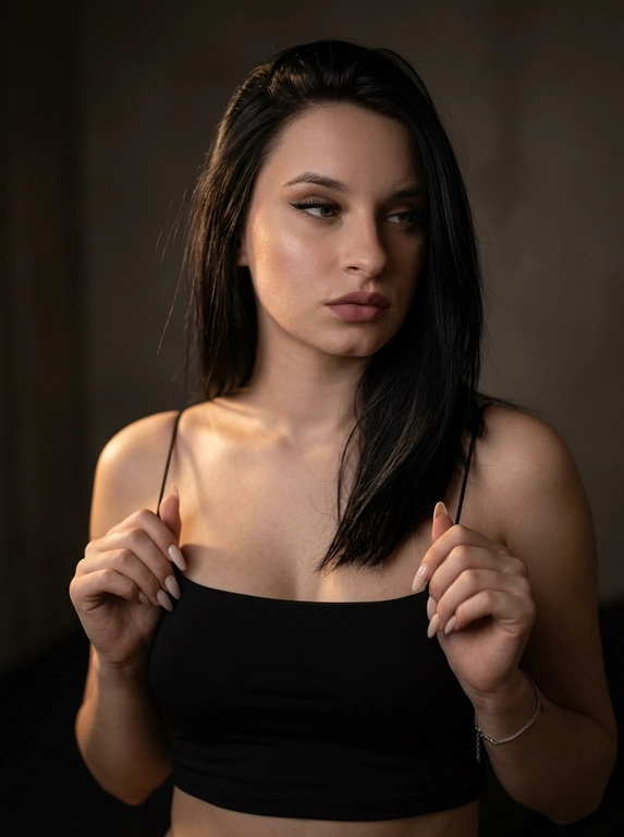 Максимально реалистичная фотография в стиле moody boudoir editorial, передающая атмосферу интимности, чувственности и приглушенного драматизма.
Крупный план по пояс (waist-up shot / close-up). женщина позирует в полумраке, придерживая руками края одежды. [Поза/взгляд/эмоция]: Плечи слегка приподняты, руки с длинным светлым маникюром сжимают тонкие бретели черного топа; голова немного повернута в сторону, взгляд задумчивый и отведен в сторону; выражение лица томное, загадочное и уверенное.
Вечерний «glam» выразительные стрелки, густо накрашенные ресницы, создающие эффект «кошачьего взгляда», мягкий контуринг лица и матовая помада цвета пыльной розы.
Облегающий черный кроп-топ на очень тонких бретелях-спагетти. Материал — матовая эластичная ткань. На запястье виден изящный серебряный браслет.
Минималистичный интерьер в темных тонах. Фон однотонный, слегка текстурированный, уходящий в глубокую тень, что создает эффект камерности.
 Низкий ключ (low-key lighting). Мягкий, теплый направленный свет падает сбоку, подчеркивая рельеф ключиц, линию плеч и текстуру кожи. Глубокие, кинематографичные тени создают объем и драматичное настроение.
 85mm f/1.4, идеальное фокусное расстояние для портрета, создающее минимальные искажения и сильное размытие фона. Очень малая глубина резкости; фокус идеально наведен на глаза и лицо модели, в то время как плечи и руки уходят в мягкое расфокусирование.
Гиперреалистичная текстура кожи: видны поры, естественный блеск на выступающих частях лица и плеч. Отсутствие «пластикового» эффекта ретуши.
 текстура ткани топа под пальцами, тонкие волоски на макушке, мягкий градиент тени на стене.
Эстетика «dark feminine energy», интимная, теплая и дорогая атмосфера (expensive moody vibe).

Какая будет прическа?: Волосы: перекинутые на одно плечо. Текстура волос гладкая и шелковистая, с отдельными тонкими прядями, подсвеченными теплым светом.