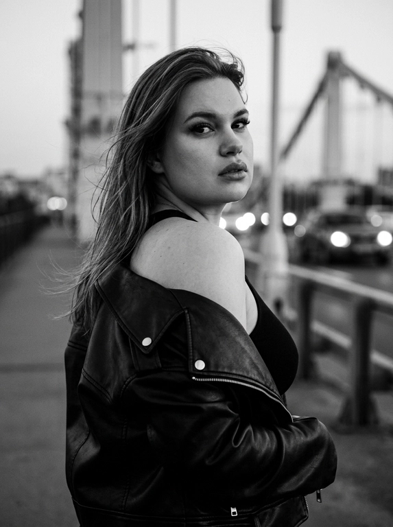 Используй внешность модели с фото. Фотореалистичное черно-белое фото в стиле модной редакционной съемки с эстетикой пленочной фотографии (зернистость, высокий контраст). Модель с волосами слегка растрепанными ветром. Она одета в черную, объемную кожаную куртку-косуху, которая небрежно спущена с левого плеча, открывая его. Под курткой надет черный облегающий топ. Поза: модель стоит вполоборота спиной к камере, но резко оборачивается и смотрит назад через плечо прямо в объектив. Выражение лица серьезное, дерзкое и чувственное, губы слегка приоткрыты, густые ресницы. Фон размыт, видны элементы конструкции моста и огни проезжающих машин вдали, создающие городскую атмосферу. Освещение естественное, подчеркивающее текстуру кожи и блеск кожи куртки. Высокое разрешение, 8k. Освещение мягкое и равномерное, без жестких теней, свет подчеркивает блеск жемчуга и страз, цвета насыщенные, но реалистичные. Высокая детализация, фотореализм, четкий фокус на лице, естественные пропорции.