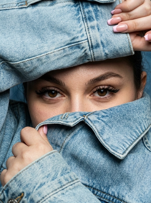 Экстремально крупный фотореалистичный fashion editorial портрет. Камера очень близко к лицу, плотный кадр, ракурс почти на уровне глаз с лёгким верхним углом. Модель не стоит прямо, а будто прячется внутри объёмной светло-голубой джинсовой куртки. Поза читается только через руки, лицо и ткань.

Весь кадр заполнен джинсой и руками. Сверху большой слой денима нависает над бровями, снизу поднятый воротник диагонально закрывает лицо до переносицы. Видны только глаза, брови, переносица и небольшой участок кожи вокруг глаз. Рот, подбородок, щёки, шея и причёска скрыты. Волосы почти не видны, максимум лёгкая тень у линии роста волос.

Одна рука сверху справа держит верхний край джинсовой куртки, частично обрезана кадром. Вторая рука снизу слева подтягивает воротник ближе к лицу. Пальцы естественно сжимают плотную ткань, создавая натяжение, складки и объём. Композиция тесная, асимметричная, с диагональными линиями ткани вокруг глаз.

Деним главный элемент кадра: светло-голубая washed denim ткань, плотная, матовая, тяжёлая, с выраженной фактурой, видимым плетением, мягкими потёртостями, заломами, массивными швами и контрастной строчкой. Джинсовая куртка выглядит натурально, объёмно и очень близко к камере.

Глаза смотрят прямо в камеру из-под бровей. Взгляд спокойный, глубокий, уверенный и слегка загадочный. Один глаз немного в тени, второй более чёткий и освещённый. Макияж глаз аккуратный: тонкая вытянутая стрелка, лёгкий cat-eye эффект, подчёркнутые ресницы, натуральные брови. В глазах живые естественные блики.

Кожа вокруг глаз реалистичная, с натуральной текстурой, мягкими порами и естественными бликами. Без пластикового сглаживания, без чрезмерной ретуши, без AI-эффекта.

Освещение мягкое, естественное, слегка контрастное, как в дорогой editorial-съёмке. Свет подчёркивает глаза, переносицу, руки, швы и складки денима. Тени глубокие, но натуральные. Цвета спокойные и реалистичные: светло-голубой деним, нейтральный тон кожи, мягкие тени.

Фон полностью отсутствует. В кадре только джинсовая ткань, руки и узкий фрагмент лица. Ultra realistic close-up, luxury denim editorial, natural film grain, shallow depth of field, soft sharpness, high detail, realistic skin texture, realistic denim texture, no CGI, no plastic skin, no over-retouching.

Negative prompt: visible hairstyle, loose hair, open face, full face, mouth visible, chin visible, neck visible, full body, standing pose, normal jacket pose, symmetrical framing, empty background, studio backdrop, glossy denim, plastic skin, AI smoothing.