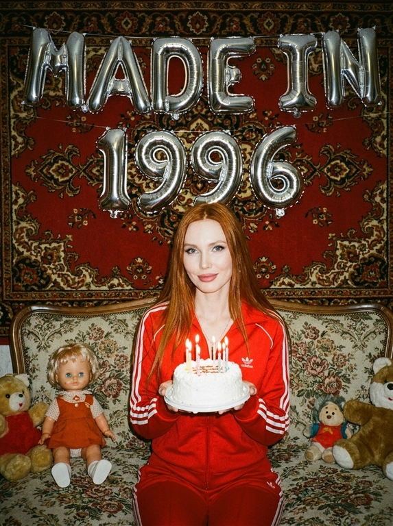 Создай крупный портрет, не меняя черты лица. На фотографии модель в стиле «лихие 90-е» в России. Модель сидит на старом диване с цветочной обивкой на фоне красного советского ковра на стене и смотрит в камеру с лёгкой улыбкой. На ковре висят большие надувные серебряные фольгированные шары в форме цифр и надпись «MADE IN». На модели классический красный спортивный костюм того времени и кроссовки. В руках маленький праздничный белый торт со свечами. Рядом на диване лежат игрушки: советские игрушки и старая советская кукла. Фото со вспышкой. Освещение мягкое, создающее атмосферу ностальгии. Высокое разрешение, реализм. Модель в олдскульном спортивном костюме сидит расслабленно, с лёгкой улыбкой. Ретро-вайб, приглушённые цвета, зернистость плёнки, детализированные текстуры ткани и шерстяного ковра.

Какая будет цифра?: Цифра 1996.