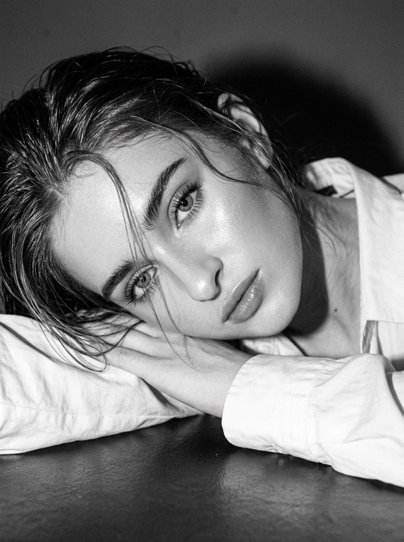 Не меняйте черты лица. Ультрареалистичный черно-белый портрет крупным планом, вертикальный, снятый с сильной прямой вспышкой на камере, создающий резкие блики и глубокие тени, ярко выраженная эстетика Y2K с более глубоким зернистостью аналоговой пленки и текстурированной монохромной цветоградаданием. Поза идентична: женщина лежит на боку, опираясь головой на сложенные руки на гладкой поверхности, тело слегка наклонено к камере, плотный кадр фокусируется на лице и плечах. Волосы, уложенные в слегка влажную редакционную текстуру с распущенными прядями, падающими по лицу точно так же, как эталон, создавая интимный естественный вид. Макияж точно в соответствии с эталоном: четкие натуральные брови, мягко затененные веки с тонкой подводкой для глаз и длинными натуральными ресницами, гладкая кожа с нежным блеском на скулах и переносице, мягко очерченные щеки, натуральные атласные губы с нежным блеском. Одежда: белая рубашка оверсайз с расслабленным воротником и видимыми складками ткани. Освещение: сильная вспышка, создающая четкие глянцевые блики на коже и прядях волос, глубокие тени и размерный контраст, высокая детализация кожи и аутентичной текстурой, ярко выраженное наложение зерна пленки Y2K, редакционная фотография красоты.