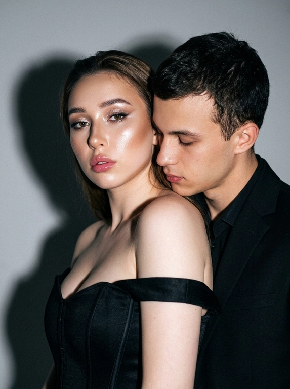 Не меняя черты лица, студийная fashion-съемка пары в минималистичном интерьере с серо-белым фоном, атмосфера гламура и интимной близости, ракурс — крупный портрет по грудь с легким поворотом корпуса, женщина на переднем плане, мужчина позади склоняется к её плечу, поза — она слегка отвернута от камеры с прямым взглядом, он касается лицом её шеи/плеча, одежда — на женщине черный корсет/бандо с открытыми плечами, на мужчине классический черный пиджак и рубашка, макияж — glam (ровный сияющий тон, сильный хайлайтер, четкий контуринг, стрелки, густые ресницы, глянцевые губы), свет — жесткая фронтальная вспышка + мягкий заполняющий свет, выраженные тени позади, эффект flash photography / editorial, холодно-нейтральная цветокоррекция с контрастом и глянцем кожи, высокая детализация, slight film grain, shallow depth of field, съемка на Canon EOS R / 50mm, f/2.0, direct flash, studio look.
