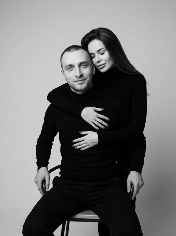 Черно-белая студийная портретная фотография в стиле минимализма. На снимке изображены мужчина и женщина, позирующие на нейтральном светлом фоне. Мужчина сидит на стуле лицом к камере, его руки расслабленно лежат на подлокотниках, взгляд направлен прямо перед собой с мягкой полуулыбкой. Женщина стоит сзади, нежно обнимая мужчину за грудь руками, а ее голова аккуратно прислонена щекой к макушке мужчины с закрытыми глазами. Оба одеты в одинаковые черные водолазки с длинными рукавами. Атмосфера кадра интимная, теплая, спокойная и эмоционально близкая. Цветовая гамма монохромная, выдержанная в контрастных черно-серых тонах с мягким студийным освещением, подчеркивающим текстуру одежды и контуры лиц.