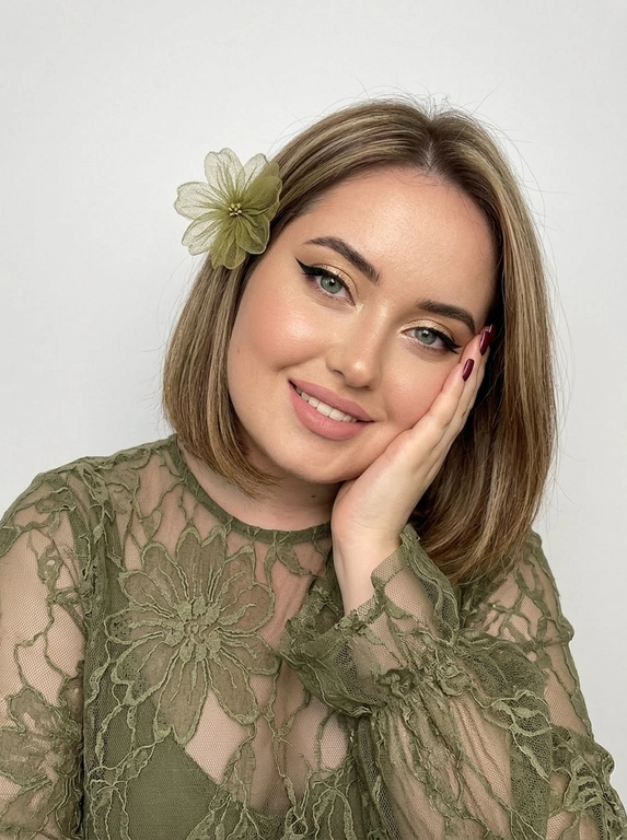 На фотографии запечатлена женщина крупным планом, позирующая для портрета. Снимок подчеркивает её ухоженную внешность и стильный образ.
Лицо и выражение:
•  Глаза:  ярко выделены макияжем. На верхних веках нанесены мерцающие золотисто-бежевые тени, дополненные четкими, длинными черными стрелками («кошачий глаз»). Ресницы густые и длинные, подчеркивающие глубину взгляда.
•  Брови: Густые, уложенные вверх (эффект ламинирования), что придает лицу современный и открытый вид.
•  Кожа: Тон кожи ровный, с нежным персиковым румянцем на скулах. Кончик носа и скулы слегка подсвечены хайлайтером для придания лицу сияния.
•  Губы: Выразительные, накрашены матовой помадой нежно-розового оттенка. Верхняя губа имеет четкий контур.
•  Поза: Женщина слегка наклонила голову вправо и подперла щеку правой рукой. Этот жест придает снимку нотку нежности и кокетства. Маникюр темно-бордовый,квадратный.
В волосах один нежный, воздушный цветок.
Одежда: На женщине надета кружевная блузка из легкой, полупрозрачной ткани.
•  Ткань украшена в крупный кружевной цветок .
•  Рукава имеют пышные воланы или оборки у запястий, что добавляет образу романтичности и женственности.
Фон и освещение:
•  Фон белый студийный
•  Освещение мягкое, направленное на лицо, что позволяет рассмотреть все детали макияжа и создает приятную, эстетичную картинку.
Общее впечатление:
Фотография представляет собой классический бьюти-портрет. Образ женщины сочетает в себе элегантность, современность и романтичность. Тщательно проработанный макияж и стильная одежда создают гармоничный и привлекательный облик.

Какого цвета будет блузка?: Оливковая 

Какого цвета будет цветок?: Оливковый