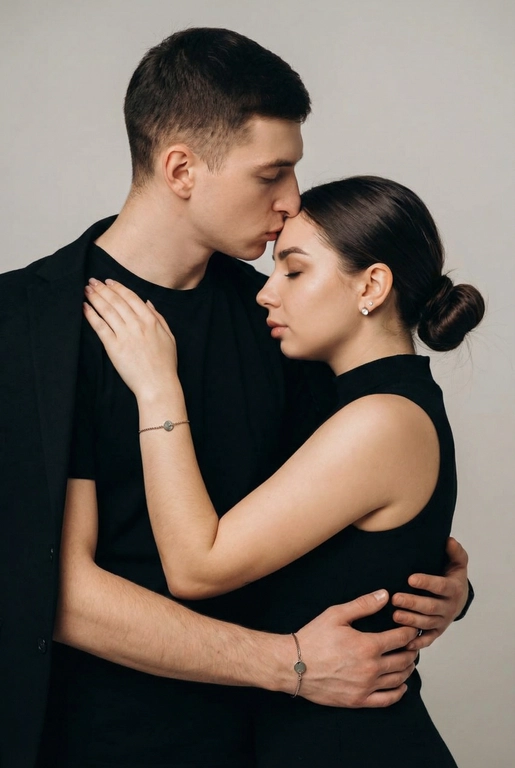   Мужчина и женщина позируют в студии в объятиях. Мужчина стоит позади женщины, обнимая её обеими руками; его тело повернуто к ней, голова слегка наклонена вниз, губы касаются её лба. Женщина немного отклоняет голову назад к его плечу, её глаза закрыты. Кадр — средний, от талии до головы, снят на уровне глаз. Композиция плотная, акцентирует линии рук и соприкосновение лиц. Женщина стоит, прямо обращённая к камере.
На ней облегающий чёрный топ без рукавов с высоким воротом, из плотной матовой ткани.
Мужчина одет в чёрную футболку и чёрную неброскую куртку. На обоих запястьях — тонкие металлические браслеты с минималистичными круглыми подвесками, а женщина носит небольшие светлые металлические серьги с гладкой поверхностью.
Обстановка минималистичная: однотонный светло-серый фон без текстуры.
Освещение — мягкий студийный свет: рассеянный фронтальный и боковой свет создаёт плавные переходы света и тени на лицах и руках. Тени мягкие, средней глубины. Контраст умеренный, без жёстких бликов. Свет равномерно распределён по фигурам, фон подсвечен мягко, без выраженных градиентов.
Съёмка на портретный объектив 85 мм, малая глубина резкости с чётким фокусом на лицах и руках; фон слегка размыт, текстуры кожи и ткани высоко детализированы. Это минималистичный студийный портрет, высокодетализированный и фотореалистичный.
Минималистичное романтическое студийное настроение, нейтральные тёплые тона кожи, низкая–средняя насыщенность, мягкое рассеянное освещение, средний контраст с нежными тенями, плавные переходы бликов, естественная передача кожи, чистый светлый фон, тонкий тональный баланс, современная мягкая редакционная цветокоррекция. Волосы женщины собраны в низкий, гладкий пучок с аккуратно уложенными прядями.