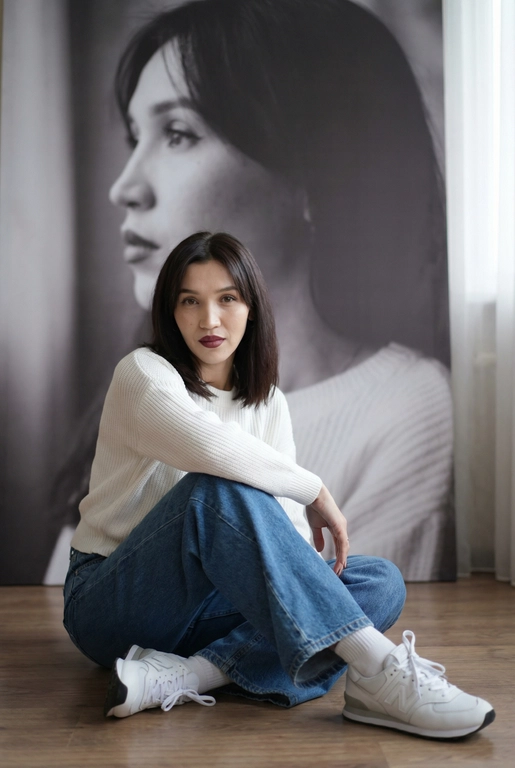   Максимально сохранить внешность, причёску и длину, как на фото.
Женщина должна сидеть на полу в изящной позе, в современном белом джемпере , в широких джинсах и белых носках и белых  кроссовках New Balance.

Её волосы как на фото
Выражение лица, как на фото, она смотрит на камеру .

Фон — тот же её профиль, в мягком крупном чёрно-белом художественном стиле, крупный план сбоку.

Композиция должна быть художественной, мягкой, реалистичной.
Создай изображение, которое выглядит как настоящая фотография.
