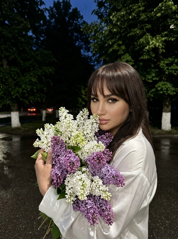 Очень крупный план, на этом фото женщина стоит в анфас к камере, обеими руками она прижимает к себе огромную пышную охапку сирени белой и фиолетовой, её голова слегка наклонена к цветам создавая ощущение что она вдыхает их аромат смотря в камеру, макияж: глаза подчеркнуты чёрными стрелками и длинными ресницами, на губах блеск, фон: действие происходит на улице в глубоких сумерках или ночью на заднем плане видны тёмные массивные силуэты деревьев с густой майской листвой внизу выкрашенные в белый цвет земля под ногами выглядит как тёмный асфальт после дождя на заднем плане едва заметные красные блики возможно огни далёких машин, ракурс: съёмка ведётся со стороны чуть сбоку на уровне глаз, освещение: слабое ночное освещение, фотореализм высокая детализация фото снято на камеру iPhone 17 Pro эффект живого кадра лёгкий цифровой шум зернистость мягкая резкость высокая детализация, без вспышки.

Какая будет одежда?: Одета в шёлковую белую рубашку
