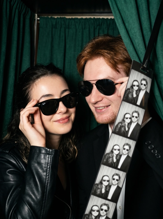 Крупный план (close-up) эффектной пары — женщины и  мужчины (не меняй лица), позирующих внутри тесной винтажной фотокабины. Они сидят вплотную друг к другу, создавая атмосферу ночного города, свидания или светской вечеринки. Композиция плотная и динамичная.

Одежда и стиль:
*   Женщина (слева): В классическом черном пиджаке. Одной рукой она кокетливо поправляет свои черные солнцезащитные очки. У нее безупречный вечерний макияж с влажным блеском на губах.
*   Мужчина (справа): Он одет в строгий черный пиджак, на нем также надеты классические черные солнцезащитные очки (Wayfarer или авиаторы), которые он слегка придерживает рукой.
*   Образ пары выдержан в едином стиле «Total Black» и эстетике «Old Money» или «Secret Agent».

Окружение и детали:
Действие происходит внутри аутентичной фотокабины. Справа на переднем плане видна свисающая длинная лента готовых черно-белых фотоснимков (photobooth film strip), на которых запечатлена эта же пара в разных позах. Фон темный, с тяжелыми складками занавески, что подчеркивает интимность момента.

Стиль и освещение:
Освещение имитирует резкую вспышку фотокабины — яркий фронтальный свет, создающий глубокие тени и подчеркивающий текстуру кожи и блеск аксессуаров. Цветовая палитра приглушенная, монохромная с теплыми телесными оттенками. Атмосфера «Nightlife Luxury».

Высочайшая детализация, акцент на блики в очках и текстуру ткани пиджаков, реалистичные черты лиц, кинематографичное качество, гиперреализм, 8k.