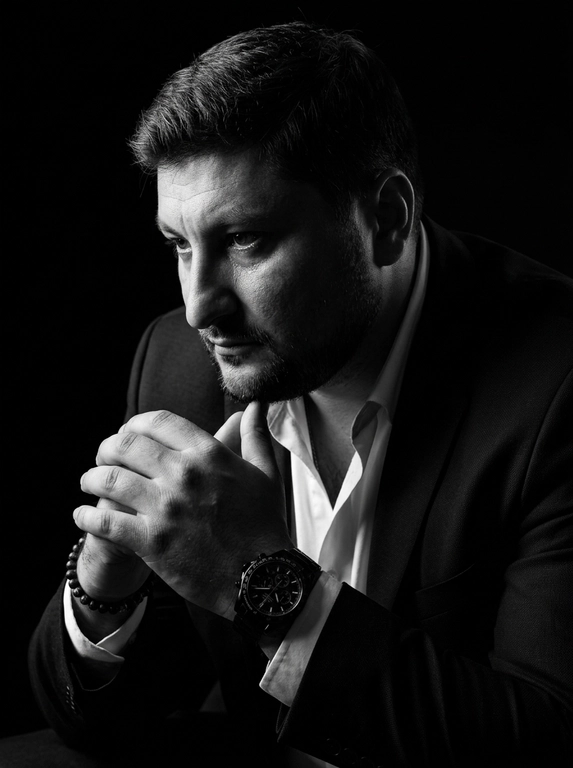 A hyper-realistic 8K cinematic portrait of a uploaded person,
captured in a dramatic Chiaroscuro style. The subject is seated at a
three-quarter angle, leaning slightly forward with a relaxed yet
commanding posture. His face is turned away from the lens, with
one side of his face clearly illuminated while the other is
completely lost in deep, velvety black shadows. His expression is
contemplative, not looking at the camera. The pose is precise: his
hands are clasped together near his chest; one hand is held
straight with fingers slightly tilted, while the other hand is
positioned so only the fingers are visible, resting over the first. He
wears a luxury black chronograph watch with a detailed metal link
bracelet, and a beaded bracelet. His attire is a sharp
black suit jacket over a white dress shirt with the top buttons
open, showing refined fabric textures. The background is a solid,
seamless black. The lighting is sharp and directional, highlighting
the intricate details of his hair,
the moisture in his eyes, and the glint off the watch face.
Cinematic black-and-white color grade with rich contrast and
smooth, natural skin transitions. Shot on 85mm lens, f/1.8, high-
resolution commercial photography