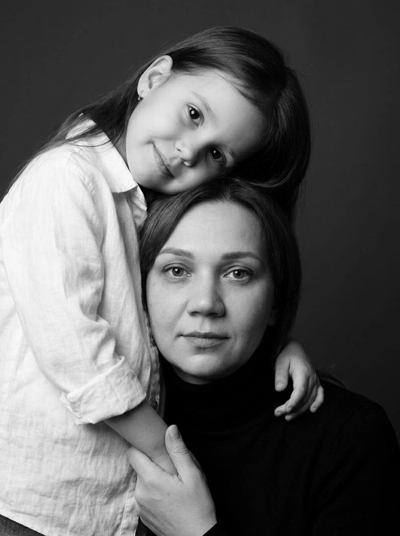 Родитель и ребёнок с внешностью как на приложенных фото, художественная семейная студийная фотосессия. Композиция — вертикальный портрет, крупный план лиц. Родитель внизу смотрит прямо в камеру, лицо фронтально. Ребёнок сверху, голова повернута набок, щека лежит на голове родителя, смотрит в камеру с нежной улыбкой. Рука ребёнка обнимает родителя за шею, видна в кадре, родитель держит руку ребёнка своей рукой.
Одежда: ребёнок в белой льняной рубашке, родитель в чёрной водолазке. 
Фон — тёмно-серый почти чёрный однотонный студийный, создаёт контраст. Освещение — мягкое боковое студийное, создаёт объём и глубину.
Чёрно-белая художественная фотография с выразительными контрастами. Атмосфера нежная, интимная, спокойная.