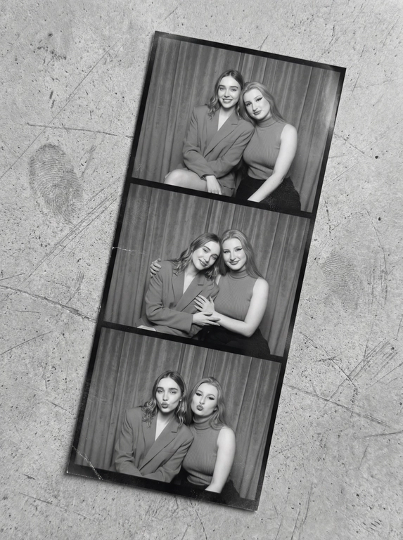   A vertical black-and-white photostrip curves diagonally across a matte, slightly scratched concrete surface, its subtle texture contrasting softly against the monochrome tones. Inside the booth, two women reside against heavy velvet curtains in deep muted gray, their backdrop textured and rich. The first frame shows them seated upright, poised yet casual, one in a wide-shouldered blazer dress, the other in a sleeveless ribbed turtleneck. Next, they lean affectionately into one another, sharing warmth and relaxed intimacy, hands gently resting or lightly touching. The final shot captures playful exaggeration: both offering dramatic, sultry pouts with slight head tilts. The strip carries the signatures of authentic photobooth prints—visible fingerprint smudges, faint creases softly bending the paper, and a delicate graininess overlaying each frame. Gentle iPhone flash reflections flicker across the surface, with shadows molding softly around their silhouettes. The whole composition tilts naturally as if casually grasped, evoking a candid, spontaneous vintage charm—contemporary photobooth snapshot, shot on iPhone.