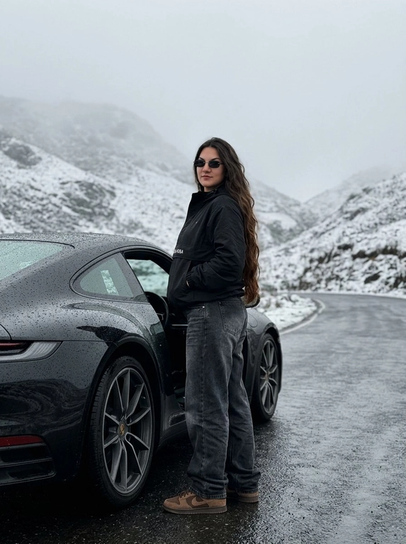 Не менять черты лица человека с исходного фото. Человек рядом с стоит рядом с машиной, машина на горной дороге, дорога мокрая от тумана, машина в каплях от тумана. Чёрный анорак Planta Rosa, маленькие чёрные солнцезащитные очки без оправы, мешковатые серо-чёрные джинсы с низкой посадкой, длинные, коричневые замшевые кроссовки Nike Dunk, руки в карманах. Машина без номеров. Фон — гиперреалистичные заснеженные холмы Мегхалайи, без зелени, туманные горы, пасмурный мягкий дневной свет, без размытия, без изменения лица или позы, без дополнительных объектов. Фото снято на iPhone 17 Pro, без фильтров на лице, без ретуши, перспектива снизу, формат 9:16, машина как на исходном фото, фон не размывать, притемнённая экспозиция, открывает дверь машины, стоит спиной, смотрит в камеру, будто сейчас сядет за руль.


Какая будет машина?: Porsche 911 самой последней модели, цвет — чёрный.