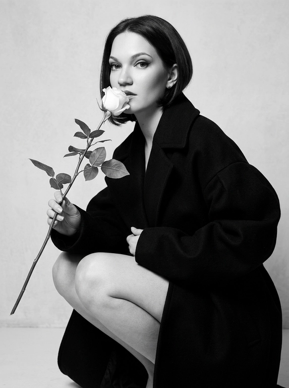 Высоко модная черно-белая редакционная фотография женщины в эффектной позе. Она грациозно присела на корточки, накинув на плечи объемное структурированное черное пальто, оставляя ноги открытыми.
Держит в руке белую розу с длинным стеблем и листьями, поднося к губам бутон. Изящный, минималистичный стиль. Сильное направленное студийное освещение, создающее глубокие тени и высокий контраст. Изысканный макияж с четкой подводкой для глаз и блеском на губах. Чистый фон, эстетика роскошного журнала, стиль Vogue, сверхчеткая, высококонтрастная монохромная фотография. Не изменяйте и не модифицируйте структуру лица, тон кожи или пропорции тела модели.
Только освещение, фон и застывшие текстуры должны быть переосмыслены в контексте задания. Сверхчеткое изображение, высокий динамический диапазон, студийная четкость, реалистичная текстура кожи, естественные поры, едва заметная зернистость пленки, редакционная цветокоррекция, съемка для печати.

Какая будет прическа?: Не изменять мою прическу.