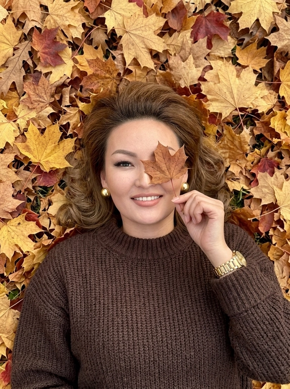 Максимально фотореалистичная фотография, снятая на iPhone. Женщина лежит на спине на плотном ковре из сухих осенних кленовых листьев золотисто-жёлтых, охристых, медных и коричневых оттенков. ракурс: камера находится сверху над женщиной, но не строго вертикально над центром лица — фотограф расположен немного со стороны её корпуса, поэтому кадр идёт сверху вниз под лёгким диагональным углом. Голова находится ближе к верхней части кадра, корпус уходит вниз, лицо и верх тела имеют естественную перспективу. Кадр достаточно близкий — женщина занимает почти всю фотографию, видна примерно от макушки до середины груди, по краям остаётся небольшое количество осенних листьев. Женщина лежит расслабленно, голова немного повернута в сторону, лицо не строго анфас, подбородок слегка развёрнут вбок, при этом глаза смотрят прямо вверх в объектив. Одна рука поднята перед лицом и согнута в локте, женщина тонкими пальцами держит за черенок один сухой коричневый кленовый лист точно напротив одного глаза— лист закрывает один глаз, второй глаз полностью открыт и хорошо виден. На женщине очень объёмный тёмно-коричневый оверсайз свитер из толстой крупной вязки, свободный в плечах и корпусе, с массивным широким высоким горлом и длинными очень широкими рукавами. На поднятой руке поверх рукава свитера находятся одни золотые часы с широким металлическим браслетом, никаких дополнительных часов или браслетов. В ушах крупные эффектные круглые золотые серьги (Не кольца!). Макияж: длинные объёмные пучковые ресницы с 2D-эффектом, кожа с естественной текстурой и видимыми порами, без ретуши. Весь фон состоит только из осенних листьев, без посторонних объектов, слегка пониженная экспозиция, натуральная цветопередача, высокая детализация, фон чёткий без размытия.