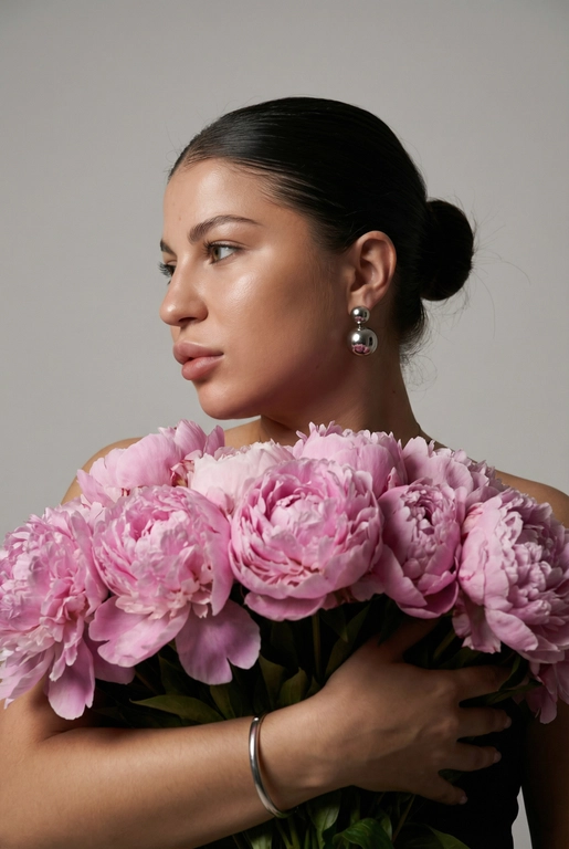 Создайте ультрареалистичный портрет, не меняя черты лица, крупным планом 3/4, с мягкой, освещённой кожей, атласной отделкой и естественным блеском, где женщина изображена в профиле. Спокойное и чувственное выражение лица, губы слегка полуоткрыты. Она носит серебряные серьги-шары и простой серебряный браслет на запястье.
Фон нейтральный, в светло-сером тоне, с тёмным, целенаправленным низким угловым освещением, которое подчёркивает контур лица, текстуру кожи и объём волос. Стиль редактирования — высокая мода, чёткий фокус 8K, реалистичная текстура, изысканная и элегантная атмосфера.


Волосы собраны в гладкий пучок

Цветы: Держит в руках огромный букет розовых пионов