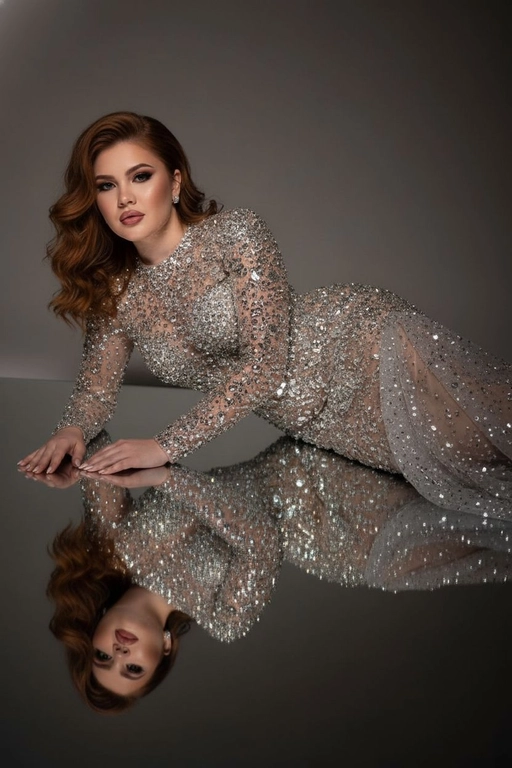 Это изысканный портрет женщины, лежащей боком на зеркальной поверхности, создавая идеальное отражение всего ее тела. На ней элегантное, облегающее платье от кутюр, полностью расшитое серебряными стразами, кристаллами и сверкающими деталями, которые роскошно ловят свет. Платье имеет длинные рукава, высокий ворот и струящийся шлейф из прозрачного тюля, усыпанного стразами на юбке, источающий гламур и изысканность. У нее длинные, волнистые волосы, которые нежно падают на ее плечи. Ее макияж безупречен с яркими темными smoky eyes и черной подводкой для глаз, длинными, объемными ресницами, безупречной кожей и пухлыми губами с изысканной, яркой красной помадой. На ней сдержанные, но элегантные серьги, дополняющие гламурный образ. Сцена драматически освещена темной виньеткой, подчеркивающей блеск платья и контраст между ее телом и отражением в зеркале. Минималистичный фон в нейтральных тонах ещё больше подчёркивает харизму модели и богатство деталей наряда. Общая атмосфера создаёт ощущение роскоши, элегантности и изысканности, отражая моду и высокую моду. Сохранены те же черты лица без каких-либо изменений, тот же тон кожи и цвет волос. Качественный маникюр, профессиональное студийное качество изображения.