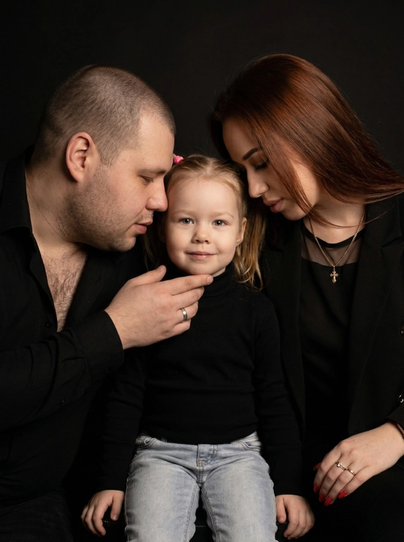 Семейный портрет из трёх человек на фоне глубокого матового черного.
Мужчина слева, слегка развернут к центру, наклонен к ребёнку, лбом почти касается его головы, ладонью поддерживает подбородок ребёнка.
Ребёнок в центре, сидит прямо, телом к камере, смотрит в объектив, в чёрной одежде с длинным рукавом.
Женщина справа, чуть повернута к ребёнку, голова наклонена вниз, лоб почти касается головы ребёнка.
Освещение мягкое, рассеянное, один источник света спереди и сверху, создаёт плавные тени и мягко выделяет лица на фоне.
Тона глубокие, нейтрально-тёплые, фон и одежда остаются темноте.