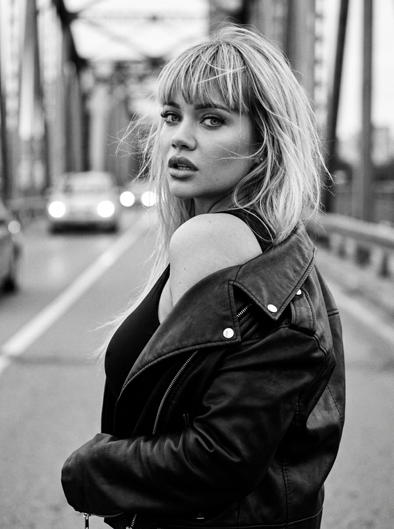Используй внешность модели с фото. Фотореалистичное черно-белое фото в стиле модной редакционной съемки с эстетикой пленочной фотографии (зернистость, высокий контраст). Модель с волосами слегка растрепанными ветром. Она одета в черную, объемную кожаную куртку-косуху, которая небрежно спущена с левого плеча, открывая его. Под курткой надет черный облегающий топ. Поза: модель стоит вполоборота спиной к камере, но резко оборачивается и смотрит назад через плечо прямо в объектив. Выражение лица серьезное, дерзкое и чувственное, губы слегка приоткрыты, густые ресницы. Фон размыт, видны элементы конструкции моста и огни проезжающих машин вдали, создающие городскую атмосферу. Освещение естественное, подчеркивающее текстуру кожи и блеск кожи куртки. Высокое разрешение, 8k. Освещение мягкое и равномерное, без жестких теней, свет подчеркивает блеск жемчуга и страз, цвета насыщенные, но реалистичные. Высокая детализация, фотореализм, четкий фокус на лице, естественные пропорции.
