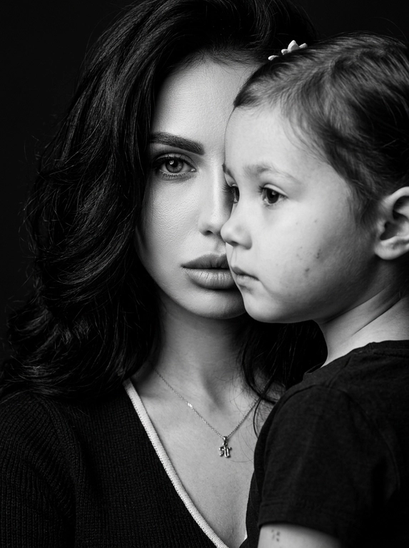 Чёрно-белый фотореалистичный студийный beauty-fashion портрет родителя и ребёнка с представленных фото. Крупный плотный кадр по плечи, без свободного пространства. Ребёнок находится на переднем плане, ближе к камере, в профиль, расположен сбоку и частично перекрывает лицо родителя. Лицо ребёнка закрывает примерно половину лица родителя, создавая сильный эффект визуального перекрытия. Родитель находится позади ребёнка, строго по центру кадра; у родителя виден только один глаз, направленный прямо в камеру, вторая часть лица скрыта лицом ребёнка. Лица расположены максимально близко, почти соприкасаются, но без явного физического действия. Ребёнок смотрит в сторону, выражение спокойное, сдержанное и закрытое. Родитель смотрит прямо в камеру одним открытым глазом, взгляд холодный, спокойный и пронизывающий. Атмосфера high-fashion editorial: минимальная эмоциональность, люксовая отстранённость, скрытое напряжение и холодная близость. Фон тёмный, чистый, студийный, без деталей. Жёсткое студийное освещение, высокий контраст, глубокие тени, выразительная светотень на лицах, натуральная текстура кожи, sharp focus, dramatic black and white photography, editorial beauty portrait, без улыбок, без лишних предметов, без текста и логотипов.