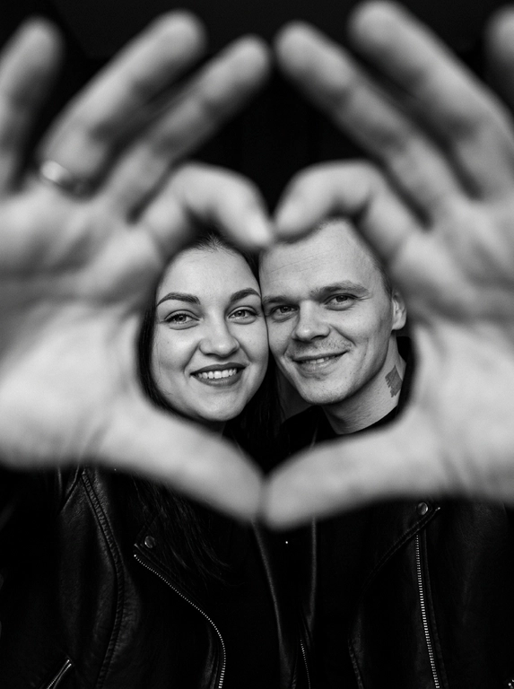Ultra-realistic editorial photography of a smiling couple in a romantic and cinematic composition. The couple is centered in the background of the image, looking directly at the camera with a spontaneous, happy, and natural expression. In the foreground, extremely close to the lens and slightly out of focus, the couple’s hands form a heart shape perfectly framing their faces.

The composition creates strong visual depth, with sharp focus on the eyes and faces inside the heart formed by the hands. The hands occupy a large portion of the foreground, artistically blurred around the edges, creating a sophisticated cinematic effect.

Intimate, emotional, and elegant aesthetic inspired by modern perfume campaigns and premium lifestyle editorials. Soft studio lighting with delicate contrast and natural shadows. Minimalistic dark blurred background.

The couple wears minimalistic black leather jackets and sophisticated dark clothing. Genuine expressions, strong emotional connection, contemporary romantic atmosphere. Hyper-realistic skin texture, visible pores, detailed hair strands, authentic human appearance.

Black and white photography, rich tones, deep blacks, and soft highlights. 85mm lens, shallow depth of field, cinematic portrait photography, luxury editorial style, ultra detailed, hyper realistic, 8K.