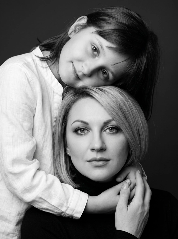 Родитель и ребёнок с внешностью как на приложенных фото, художественная семейная студийная фотосессия. Композиция — вертикальный портрет, крупный план лиц. Родитель внизу смотрит прямо в камеру, лицо фронтально. Ребёнок сверху, голова повернута набок, щека лежит на голове родителя, смотрит в камеру с нежной улыбкой. Рука ребёнка обнимает родителя за шею, видна в кадре, родитель держит руку ребёнка своей рукой.
Одежда: ребёнок в белой льняной рубашке, родитель в чёрной водолазке. 
Фон — тёмно-серый почти чёрный однотонный студийный, создаёт контраст. Освещение — мягкое боковое студийное, создаёт объём и глубину.
Чёрно-белая художественная фотография с выразительными контрастами. Атмосфера нежная, интимная, спокойная.