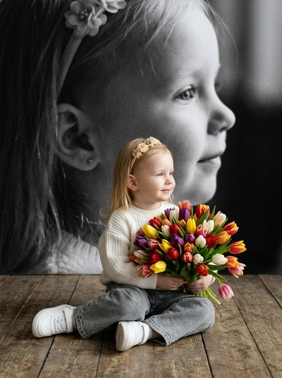 Создай портрет, не меняя черты лица, и превратите его в художественный портрет.
Ребенок сидит на полу в изящной позе, в белой кофте, мягких свободных серых джинсах и белых спортивных кроссовках. Ребенок держит в руках огромный букет ярких разноцветных тюльпанов.
Выражение лица — лёгкая улыбка, ребенок смотрит в сторону. 
Фон — тот же его профиль, в мягком крупном чёрно-белом художественном стиле, крупный план сбоку.
Композиция должна быть художественной, мягкой, реалистичной.
Создай изображение, которое выглядит как настоящая фотография. 
Не меняй черты лица.