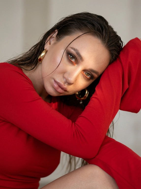 Создай не меняя черты лица. Портрет женщины крупным планом. Она склонила голову набок, опираясь на согнутую руку, что создает угловатые линии в ее позе. Ее взгляд прямой, притягивающий зрителя своей интенсивностью и уязвимостью. Ее волосы влажные, пару прядей падают на лицо.
На ней приталенный ярко-красный топ с невероятно длинными рукавами широкими на концах, сшитый из плотной матовой ткани, которая слегка облегает ее фигуру. Наряд минималистичен, но в то же время изыскан.
Освещение мягкое, но направленное, идет сверху и немного сбоку, отбрасывая нежные тени на ее лицо и подчеркивая черты ее лица.
Макияж: глаза: тени растушевка, Смоки айс, коричневый с черным цветом растушевано с дымкой, внутри слизистой прокрашено ярким оранжевым карандашом и с низу растушеван, глаза выделяются. Ее кожа плавно рассеивает свет, хайлайтер на скулах, нос и под бровью. Брови естественные, прочесаны, ресницы прокрашенные. Губы с блеском и естественную текстуру для фотореализма. Серьги объемные золотые кольца.
Свет мягко подчеркивает объем, создавая кинематографичную атмосферу. Нейтрально-светлый размытый фон усиливает концентрацию на лице. Солнечный свет частично освещает лицо. Образ мечтательный и нежный, фотореалистичный, текстурная детализация 8K. Фон светлый студийный.