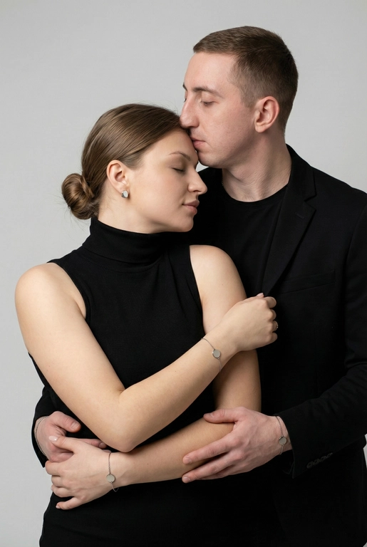   Мужчина и женщина позируют в студии в объятиях. Мужчина стоит позади женщины, обнимая её обеими руками; его тело повернуто к ней, голова слегка наклонена вниз, губы касаются её лба. Женщина немного отклоняет голову назад к его плечу, её глаза закрыты. Кадр — средний, от талии до головы, снят на уровне глаз. Композиция плотная, акцентирует линии рук и соприкосновение лиц. Женщина стоит, прямо обращённая к камере.
На ней облегающий чёрный топ без рукавов с высоким воротом, из плотной матовой ткани.
Мужчина одет в чёрную футболку и чёрную неброскую куртку. На обоих запястьях — тонкие металлические браслеты с минималистичными круглыми подвесками, а женщина носит небольшие светлые металлические серьги с гладкой поверхностью.
Обстановка минималистичная: однотонный светло-серый фон без текстуры.
Освещение — мягкий студийный свет: рассеянный фронтальный и боковой свет создаёт плавные переходы света и тени на лицах и руках. Тени мягкие, средней глубины. Контраст умеренный, без жёстких бликов. Свет равномерно распределён по фигурам, фон подсвечен мягко, без выраженных градиентов.
Съёмка на портретный объектив 85 мм, малая глубина резкости с чётким фокусом на лицах и руках; фон слегка размыт, текстуры кожи и ткани высоко детализированы. Это минималистичный студийный портрет, высокодетализированный и фотореалистичный.
Минималистичное романтическое студийное настроение, нейтральные тёплые тона кожи, низкая–средняя насыщенность, мягкое рассеянное освещение, средний контраст с нежными тенями, плавные переходы бликов, естественная передача кожи, чистый светлый фон, тонкий тональный баланс, современная мягкая редакционная цветокоррекция. Волосы женщины собраны в низкий, гладкий пучок с аккуратно уложенными прядями.
