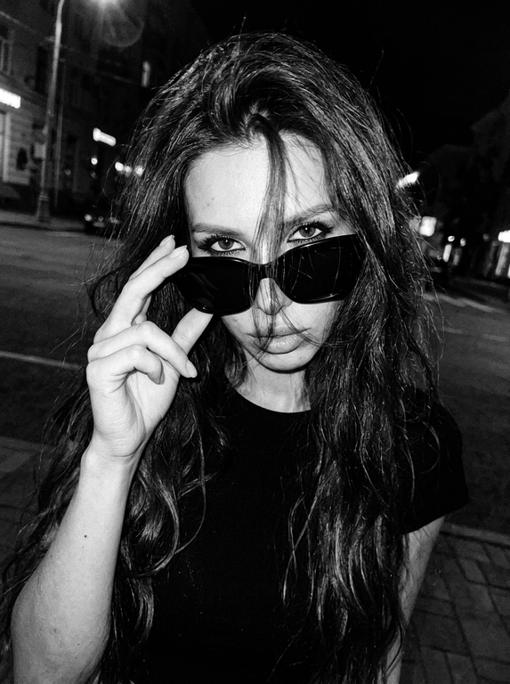 Интригующий, нуарный lifestyle момент; эстетика «messy chic» и ночного города; зернистое черно-белое фото с высоким контрастом, передающее атмосферу скрытности и дерзости.

Крупный план (extreme close-up), голландский угол (наклон камеры); имитация снимка на пленочную камеру 35mm с характерным цифровым шумом и виньеткой.

Модель игриво подносит руку к лицу, частично закрывая его пальцами; кончики пальцев касаются оправы очков или прядей волос.

Динамичный наклон головы в сторону; плечо слегка приподнято, создавая ощущение случайного, небрежного кадра «в моменте».

Загадочный, прямой взгляд в объектив сквозь линзы очков; губы слегка расслаблены; акцент на один глаз, видимый сквозь пряди волос.

Растрепанные волосы; тонкие пряди хаотично спадают на лицо и очки, создавая многослойность и текстуру.

Черный топ; акцент на аксессуары — крупные очки в массивной черной оправе.

Жесткий направленный свет (harsh flash) в темноте, создающий глубокие тени; эффект вспышки «в лоб», который выбеливает кожу и подчеркивает блеск очков и ногтей.

Выраженное пленочное зерно (film grain), легкий расфокус на заднем плане, естественные блики на линзах очков, детальная текстура кожи с сохранением естественных пор под слоем контрастных теней.

Photorealistic cinematic editorial, high-contrast black and white photography, raw aesthetic, 90s grunge vibe.  

Без: ИИ-эффектов, пластиковой кожи, пересветов, HDR, неестественных цветов. Все материалы (волосы, металл, ткань) выглядят максимально достоверно.