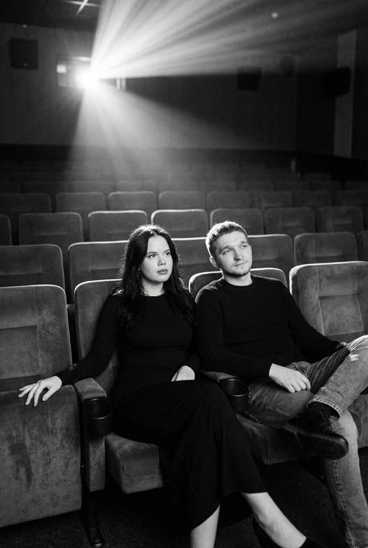  Кинематографичный чёрно-белый портрет пары в пустом кинозале.
Женщина и мужчина сидят рядом в мягких креслах кинотеатра.
кадр с глубоким пространством и пустыми рядами кресел позади них. Женщина черном длинном обтягивающем платье, поза женственная и спокойная, одна рука на подлокотнике.
Мужчина в черном свитере и в джинсах, сидит расслабленно.
Над ними яркий луч проектора, и выразительные блики.