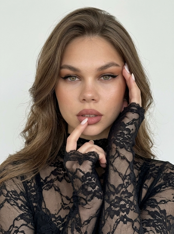 Лёгкий макияж с акцентом на глаза: чёрные cat-eye стрелки на верхнем веке, нюдовая матовая помада оттенка пыльной розы, очерченная карандашом для губ на тон темнее, влажный хайлайтер. Кожа загорелая. Ультрареалистичная фотография, естественная текстура кожи, лёгкий шум, необработанный реалистичный вид, без фильтра красоты, без CGI, visible eye capillaries, natural skin texture with pores and micro-imperfections, depth of field, subsurface scattering, soft morning light, Tyndall effect, hyper-detailed iris, unedited RAW photo, 8K resolution. Максимально фотореалистичный студийный портрет, вертикальный формат, снято на iPhone 17 Pro, 8K.

Женщина сидит, но кадр настолько близкий, что место, на котором она сидит, бёдра и ноги полностью остаются за пределами кадра. РАКУРС ОЧЕНЬ БЛИЗКИЙ: камера прямо перед женщиной, на уровне глаз, объектив близко к лицу. В кадре только голова, плечи, руки и небольшая часть верхней части корпуса. Макушка близко к верхней границе кадра. Лицо почти фронтально, совсем немного повёрнуто в сторону, взгляд прямо в объектив, губы слегка приоткрыты. Женщина сидит расслабленно, корпус слегка подан вперёд.

Обе руки подняты к лицу очень компактно. Локти находятся близко друг к другу перед корпусом и направлены вниз, не разведены в стороны. Одна кисть поднята снизу к подбородку и губам, пальцы мягко согнуты, указательный палец касается нижней губы. Вторая кисть находится немного выше, возле противоположной щеки. Запястья практически рядом. Руки располагаются по обе стороны лица, возле щёк и висков.

На женщине тонкая чёрная полупрозрачная кружевная кофта по фигуре с высоким облегающим воротником. Всё полотно состоит из нежного ажурного кружева с мелким органичным растительным рисунком из тонких переплетающихся стеблей, небольших цветов и листьев, между которыми просвечивает кожа. Это цельное тонкое кружевное полотно, не сетка с нашитой вышивкой и не тяжёлый гипюр. Рукава экстремально длинные, закрывают запястья и почти полностью заходят на кисти до оснований пальцев. Края рукавов необработанные: кружево заканчивается непосредственно по своему рисунку, образуя тонкие ажурные выступы вокруг кистей, без манжет, подгиба и ровной строчки.

Макияж выразительный: загорелая, увлажнённая, сияющая кожа с натуральной текстурой и порами, сильный бронзер и контуринг скул, влажный хайлайтер, вытянутые чёрные кошачьи стрелки, пухлые нюдово-розовые губы с более тёмным коричнево-розовым контуром.

Позади неё ровная однотонная молочная стена: светлый мягкий молочно-белый оттенок, не серая, не серо-молочная, не бежевая, без декора и предметов. Мягкий нейтральный студийный свет, натуральный баланс белого, естественные оттенки кожи. Никакой жёлтой, золотистой или тёплой обработки. Чистая натуральная фотография без фильтров, пресетов, цветокоррекции, винтажного эффекта, HDR, ретуши, боке, пластиковой кожи и ИИ-эффекта. Маникюр молочный, миндальной формы, ровный и аккуратный.

Причёска: Очень длинные густые волосы с воздушным прикорневым объёмом и крупными мягкими волнами по длине. Передние слои плавно обрамляют лицо, несколько тонких прядей естественно лежат возле щёк.